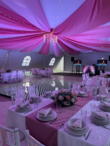 Decoración elegante de un banquete con mesas blancas y rosas bajo un toldo morado.